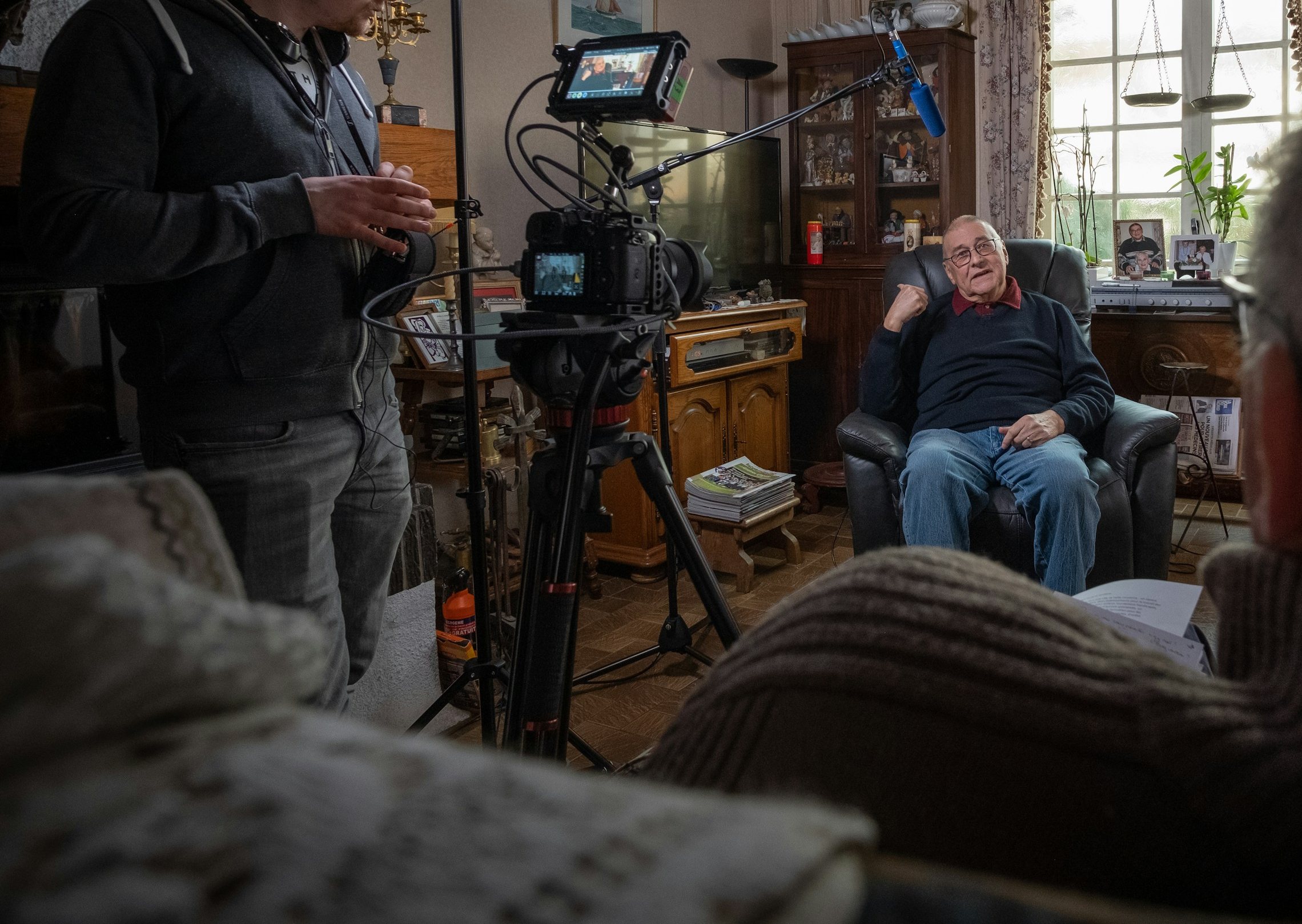 A filming day at home: an older man talks in his armchair while a camera records and the crew listens